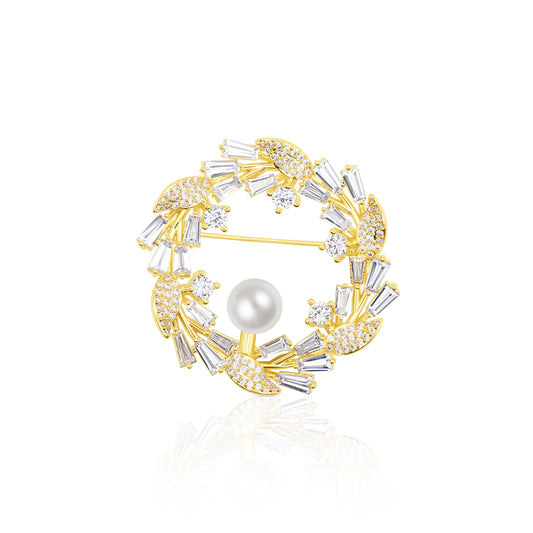 Circular Crystal Wreath Brooch with alternating clear baguette and round stones around a luminous pearl. The gold-plated setting highlights the contrast and symmetry of this festive, winter-ready accessory. Resting on a white reflective surface, it gleams with polished brilliance, making it ideal for holidays, formal events, or refined winter fashion.
