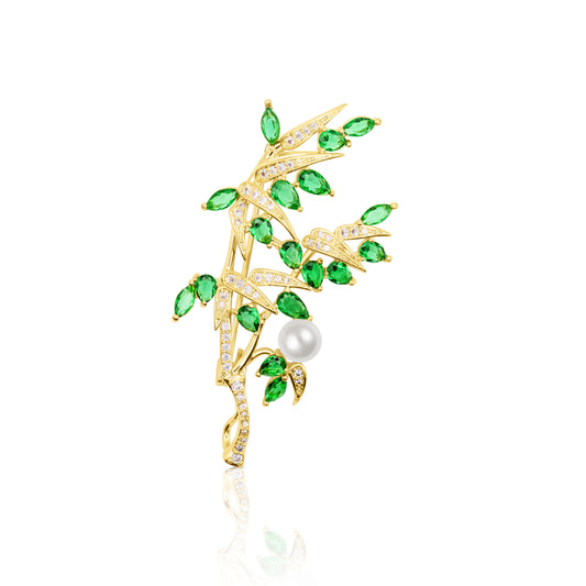 A striking bamboo-themed gold-tone brooch featuring vivid green marquise-shaped cubic zirconia arranged as leaves on curved stems accented with clear crystals. At the base of the brooch rests a luminous round faux pearl, symbolizing harmony and elegance. This nature-inspired piece exudes tranquility and is ideal for styling formal outfits or gifting as a symbol of peace and renewal.