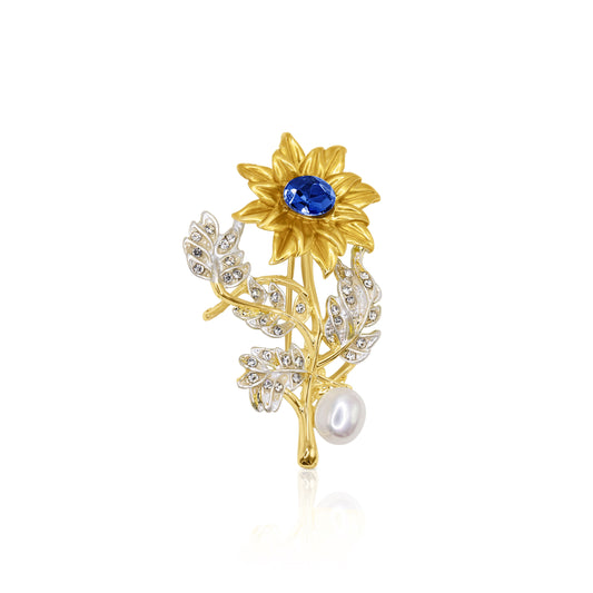 A sunflower-inspired brooch with a sapphire-blue crystal at the center, gold-tone detailing, and a pearl accent, perfect for brightening any look.